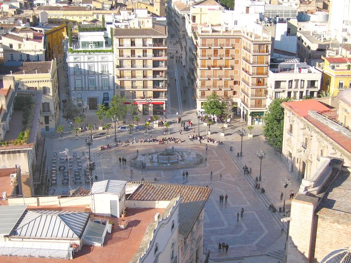 The square seen from above, with its round fountain, café tables and people walking across.