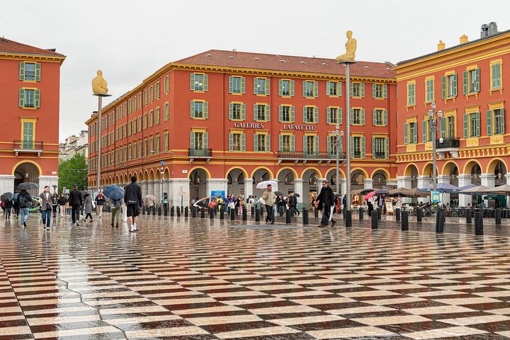 Place Masséna on a rainy day, with chequered paving, red arcaded buildings and figures on tall poles.